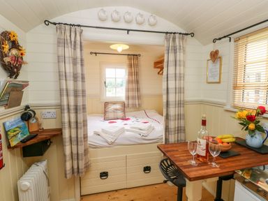 A small bedroom area with a bed, curtains, a window, a wooden table with wine bottle and glasses, and a fruit basket at Shepherds Hut - The Crook in Rosemarket near Neyland