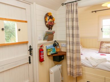 A small sleeping area with a bed, plaid curtain, wooden corner shelf, fire extinguisher, and a white door at Shepherds Hut - The Crook Rosemarket near Neyland