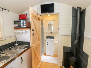 A small kitchen area with a sink microwave and toaster a wooden door leading to a bathroom with a sink and mirror and a wood burning stove in Shepherds Hut - The Crook in Rosemarket near Neyland