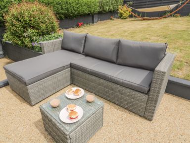 An outdoor seating area with a grey cushioned wicker sofa and a wicker table with plates of scones and cups of tea in a garden at Chi An Mor in Porth