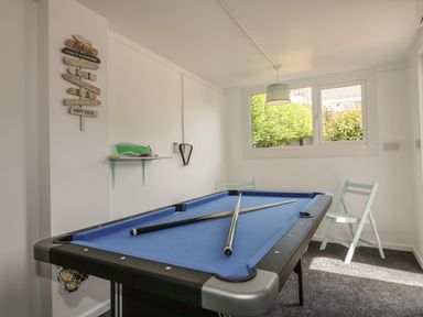 A room with a pool table with blue felt and two pool cues on it two light blue chairs a wall shelf and a window showing greenery at Chi An Mor in Porth