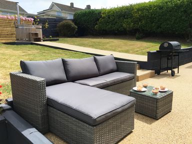 An outdoor seating area with a grey cushioned sofa and table with food and drinks a barbecue grill and a wooden hot tub in a garden at Chi An Mor in Porth