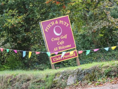 An outdoor sign advertising pitch and putt crazy golf cafe with clubs provided and colorful flags in front of trees at Phenests in Falmouth