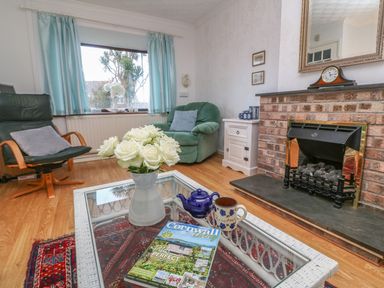 A living room with a glass coffee table with flowers cups and a magazine in front of a brick fireplace with a clock and two chairs at Phenests in Falmouth