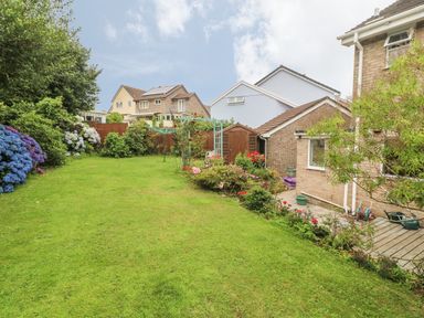 A garden with green grass and flower bushes next to a brick house and sheds at Phenests in Falmouth