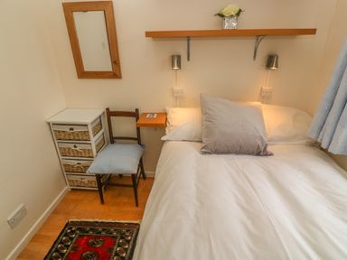 A bedroom with white bed linens a gray pillow a wooden chair with a cushion a small cabinet with baskets a mirror and a shelf with flowers at Phenests in Falmouth