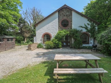 A house with arched windows surrounded by trees and plants a wooden picnic table on grass and a gravel driveway at Beggars Roost in Penhallow