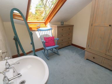 A room with a blue rocking chair with a pink cushion a wooden chest of drawers a wooden wardrobe a freestanding mirror and a white sink under a skylight window at Beggars Roost in Penhallow