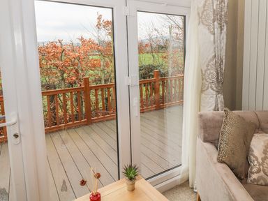 A corner of a living room with a beige sofa and pillows next to glass doors opening to a wooden deck with a railing and a view of trees and green fields at Avonal Lodge in Dunning