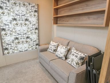 A room with a gray sofa with animal patterned cushions a window with matching animal print curtains wooden shelves and folded black chairs at Avonal Lodge 24 in Dunning