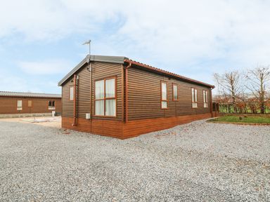 A wooden cabin on a gravel driveway with trees in the background at Avonal Lodge 24 in Dunning