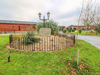 A circular fenced garden with a stone and lamp post surrounded by grass and wooden buildings at Avonal Lodge in Dunning