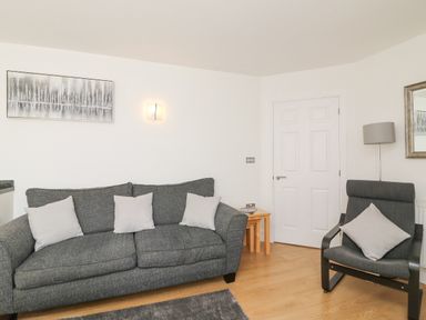 A living room with a gray sofa and armchair with white cushions a small wooden table a wall lamp and artwork at Admirals Quarter Apartment 4 in Weymouth