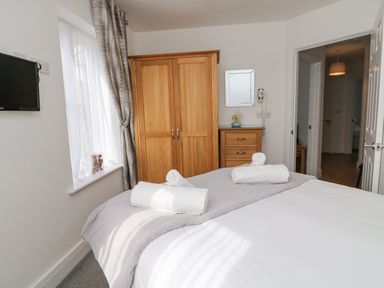A bedroom with a bed, wooden wardrobe, chest of drawers, wall mounted TV, window with curtains and a doorway at Admirals Quarter Apartment 4 in Weymouth