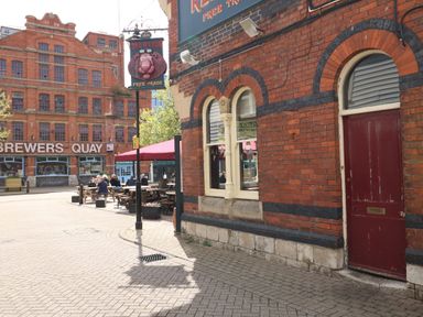 A street view with a red brick building and a pub sign reading The Red Lion with outdoor seating and another large building in the background with Brewers Quay on it