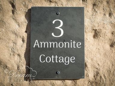A black metal house number sign with the number 3 and text Ammonite Cottage on a rough stone wall at Ammonite Cottage Fortuneswell Portland
