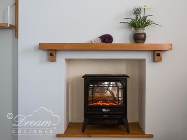 A small black electric stove heater in a recessed space with a wooden shelf above holding a vase with flowers and a small knitted item at Ammonite Cottage in Fortuneswell Portland