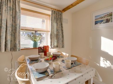 A dining table set with bowls, mugs, cutlery and fruit under a window with floral curtains at Ammonite Cottage in Fortuneswell Portland