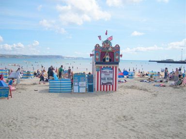 A beach scene with people swimming and relaxing on sand near a Punch and Judy show booth at Anvil House Brewers Quay Harbour