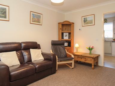 A living room with a leather sofa two cushions a wooden chair a wooden corner cabinet a wooden table with lamp and flowers and framed pictures on walls at Anvil House in Brewers Quay Harbour