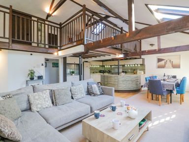 An open living space with a gray sectional sofa coffee table kitchen area and dining table under wooden loft beams at Barley Mews Brewers Quay Harbour
