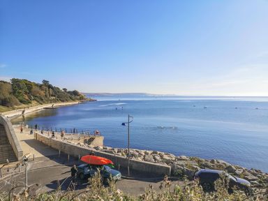 A coastal walkway with people exercising near calm sea and cars parked by rocks at Barley Mews in Brewers Quay Harbour