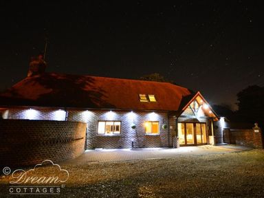 A brick cottage with exterior lights and a thatched roof at night with a gravel driveway at Baytree Lodge in Osmington