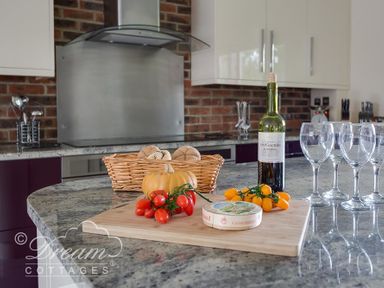 A kitchen counter with a wooden cutting board holding tomatoes cheese a small pumpkin and a bottle of wine next to four wine glasses at Baytree Lodge in Osmington