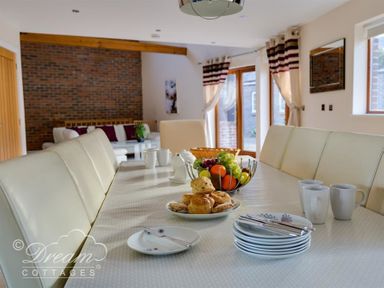 A dining table with plates cutlery mugs scones and a fruit basket in a room with a brick wall and windows at Baytree Lodge in Osmington