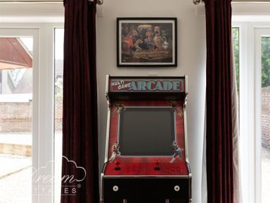 An arcade game machine between two windows with dark curtains and a framed picture above at Baytree Lodge in Osmington