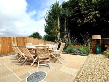 An outdoor patio with a wooden table and chairs next to a garden and wooden play structure at Baytree Lodge in Osmington