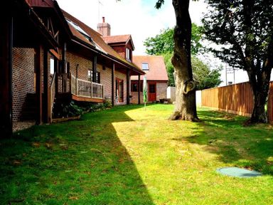 A brick house with wooden porch and large trees in a fenced green backyard at Baytree Lodge in Osmington
