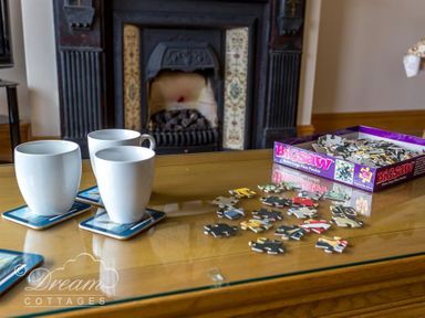 A table with three white mugs on coasters and a partially assembled jigsaw puzzle with a fireplace in the background at Beach View Apartment 2 in Greenhill