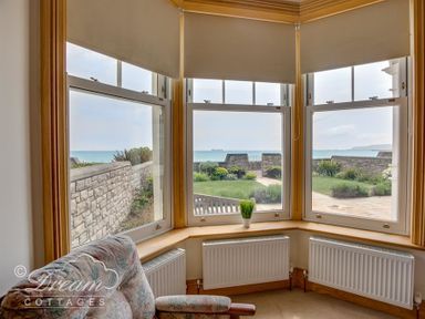 A corner bay window with roller blinds looking out to a garden and sea beyond with a floral armchair inside at Beach View Apartment 2 in Greenhill