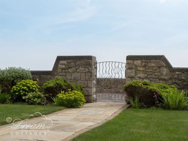 A stone pathway leading to a metal gate with fish designs set in a stone wall with bushes on both sides at Beach View Apartment 2 in Greenhill