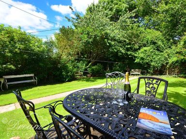 An outdoor garden area with a black metal table and chairs a bottle of champagne glasses and a travel magazine on the table benches and green trees in the background at Little Berwick Cottage Burton Bradstock