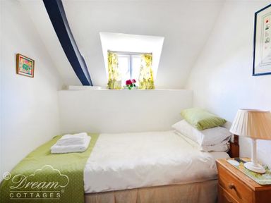 A small bedroom with a single bed a bedside table with a lamp and a window with floral curtains at Little Berwick Cottage in Burton Bradstock