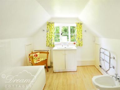 A bathroom with a bathtub a wooden chair a sink under a window with yellow floral curtains and a heated towel rail at Little Berwick Cottage in Burton Bradstock