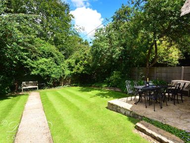 A garden with a stone patio and black metal table and chairs a grass lawn a wooden bench and trees and bushes at Little Berwick Cottage in Burton Bradstock