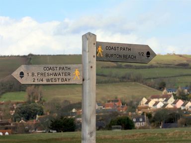 A wooden signpost showing directions for coast path to Burton Beach and West Bay in a rural area