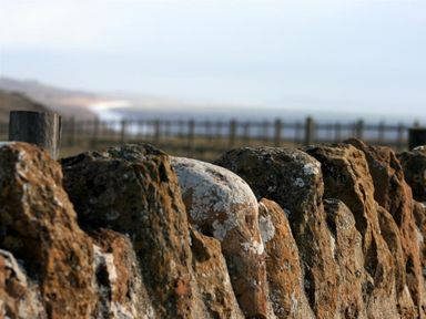 A stone wall with weathered rocks near a wooden post and a fence in the background at Little Berwick Cottage in Burton Bradstock