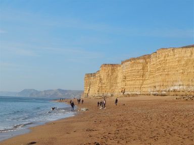 A beach with people walking near tall layered cliffs and the sea at Little Berwick Cottage in Burton Bradstock