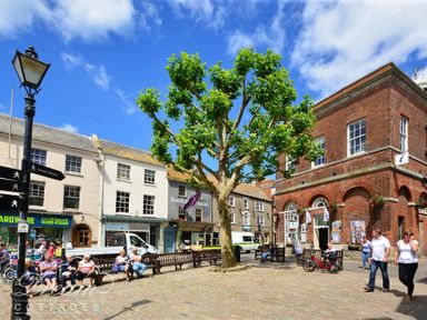 A public square with benches a tree people walking and sitting and old brick and stone buildings at Little Berwick Cottage in Burton Bradstock
