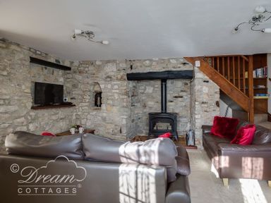 A sitting room with stone walls a wood burning stove two brown sofas with red cushions and wooden stairs at Blueberry Cottage in Sutton Poyntz