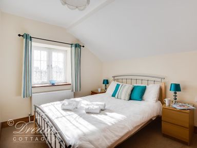 A bedroom with a double bed with white bedding and teal pillows a window with striped curtains and two wooden bedside tables with lamps at Blueberry Cottage in Sutton Poyntz