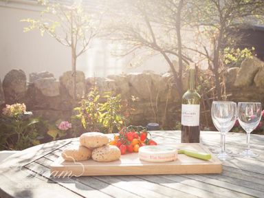 An outdoor table with bread rolls cherry tomatoes cheese a knife two empty wine glasses and a bottle of wine at Blueberry Cottage in Sutton Poyntz