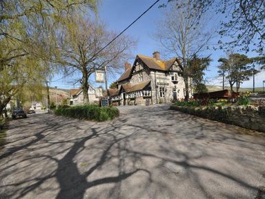 A street view with a stone building surrounded by trees and parked cars at Blueberry Cottage in Sutton Poyntz