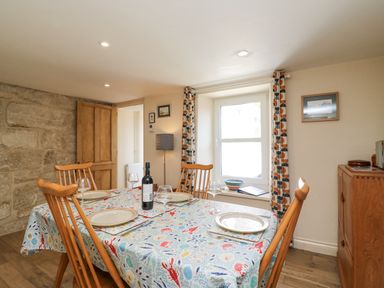 A dining room with a wooden table set with plates glasses and a bottle a window with patterned curtains and a wooden cabinet at Bowmans Cottage in Wakeham Portland