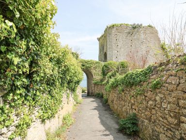 A narrow pathway between stone walls leading to an arched stone gateway and an old stone tower with greenery at Bowmans Cottage in Wakeham Portland