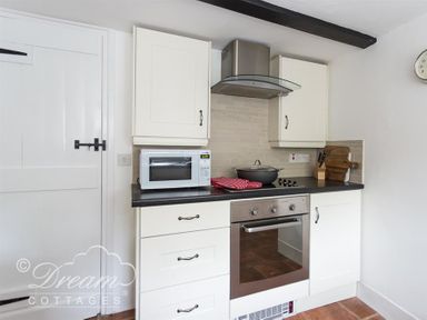 A kitchen with white cabinets a microwave a stovetop an oven and a wooden cutting board at Bramble Cottage in Burton Bradstock